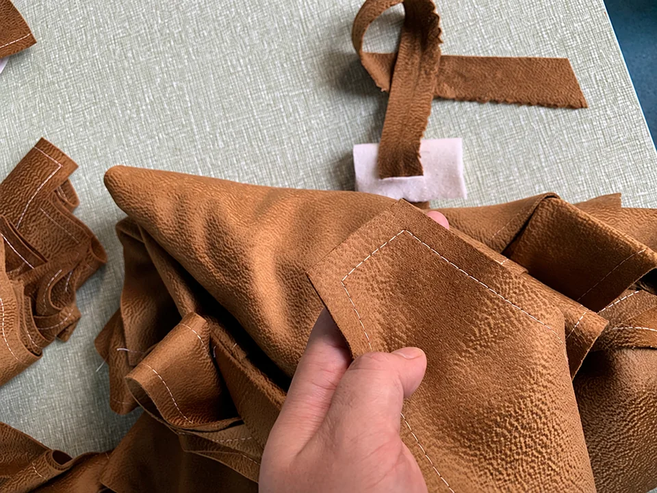 A hand holding brown fabric cut pieces with visible white preparation stitches