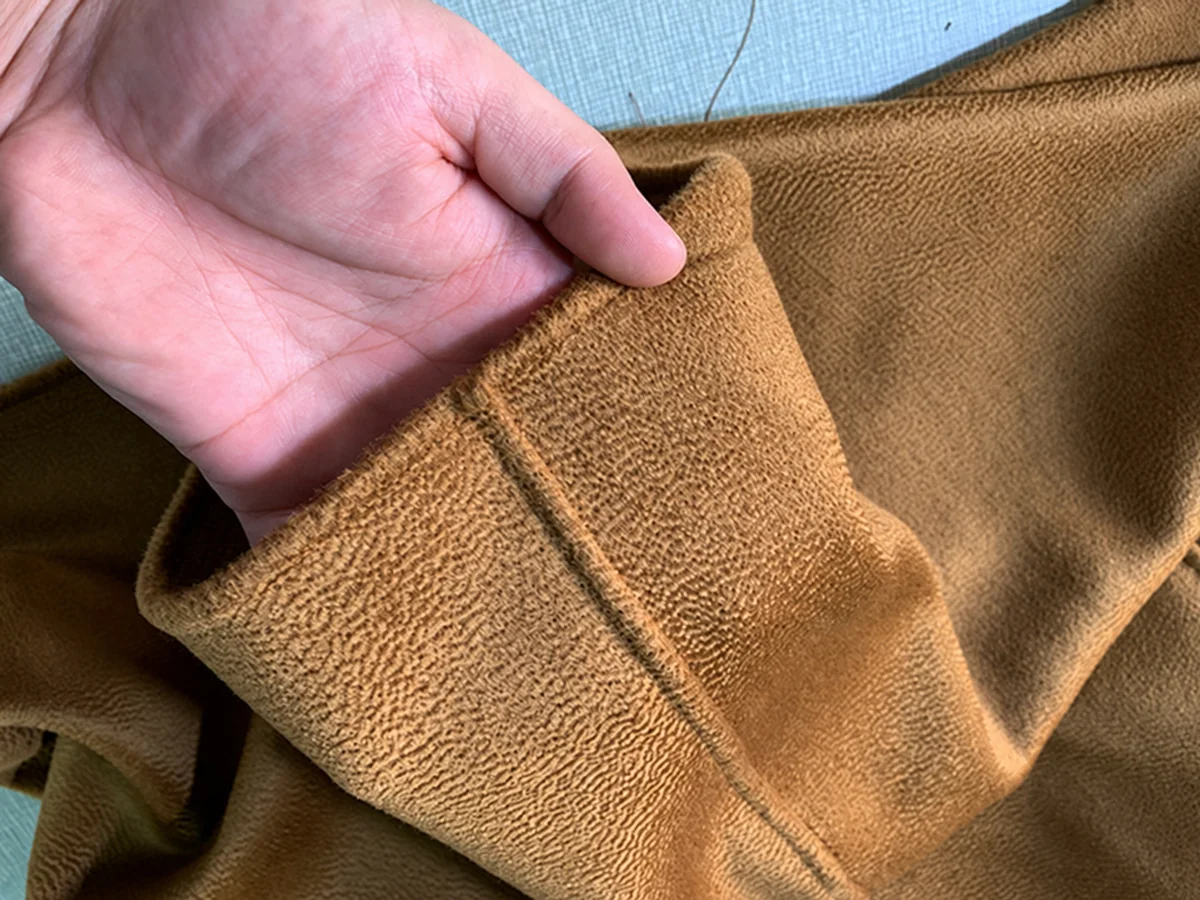 A hand holding open a camel-coloured garment edge to show its seam finish